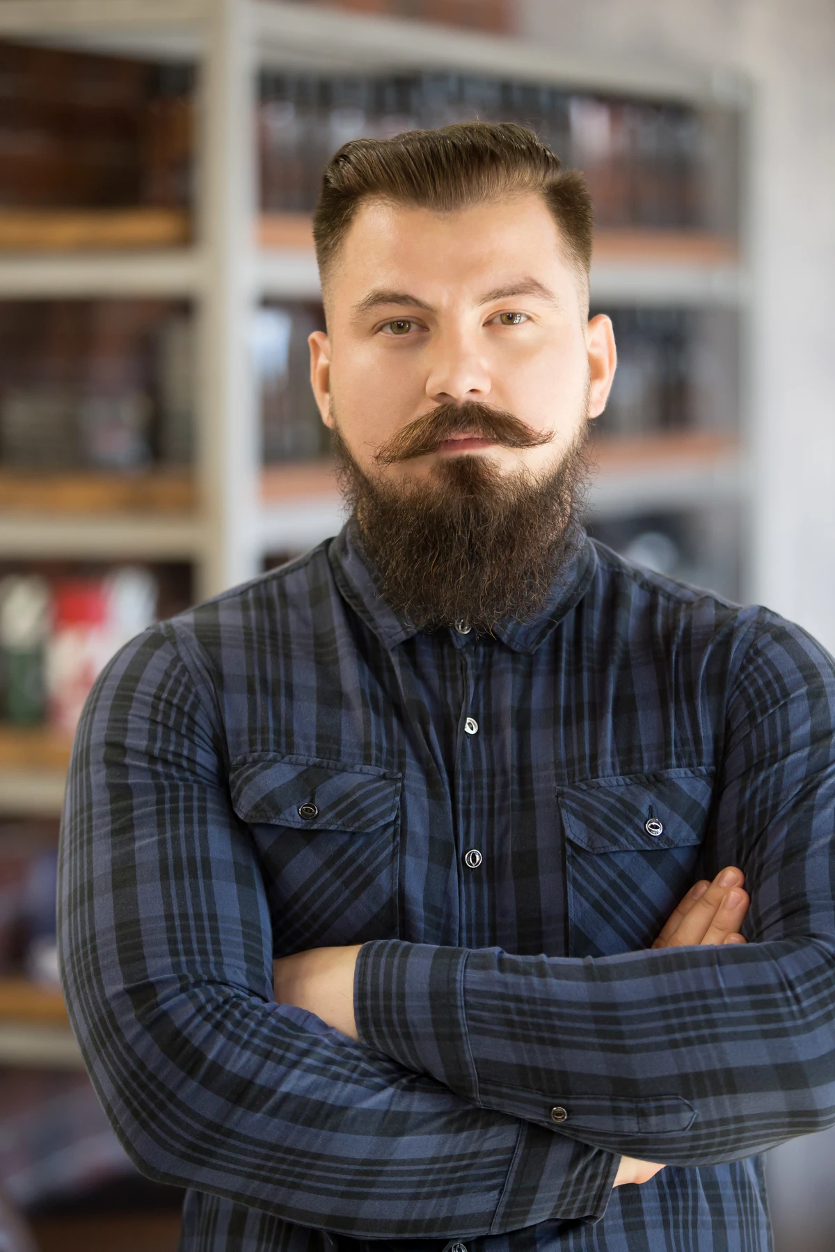 Headshot portrait of serious young man with long beard and mustache. Handsome bearded man posing with hands crossed on his chest, looking at camera, standing in his shop por Reprodução | Freepik