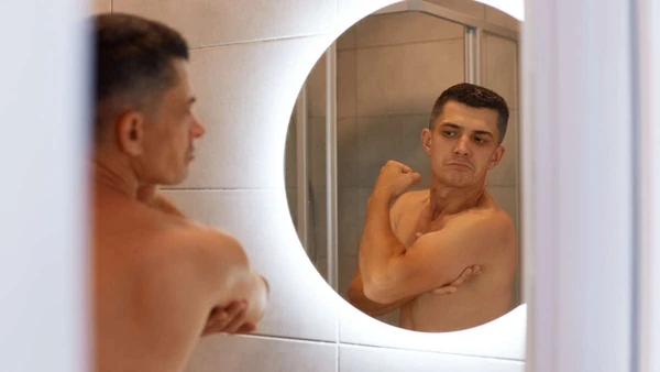 Reflection in the mirror self-confident proud male with dark hair and naked body, looking at his biceps with satisfied facial expression, posing in bathroom.