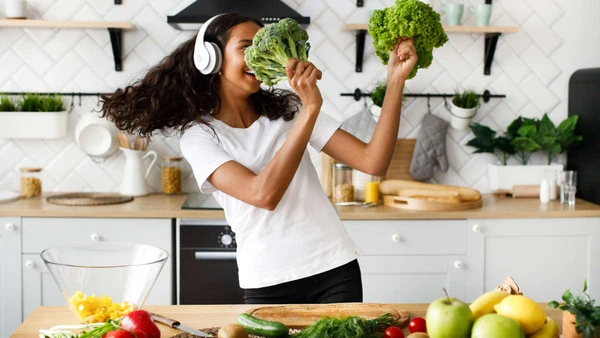 Smiled mulatto girl in big wireless headphones is dancing with salad leaves and broccoli on the modern kitchen near table full of vegetables and fruits