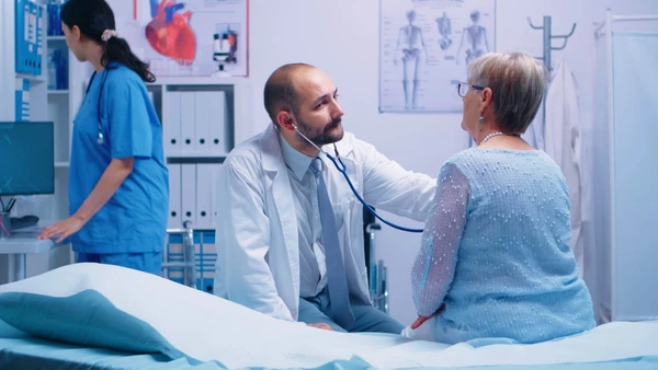 Elderly woman doctor appointment in modern private clinic while she sitts on hospital bed and the physician is listening her heart beat. Health care medicine treatment, specialist consultation