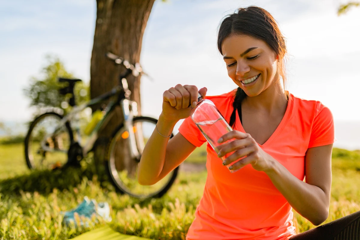smiling beautiful woman drinking water in bottle doing sports in morning in park nature doing yoga on mat, pink fitness outfit, happy healthy lifestyle, music in earphones, bicycle on background por Reprodução | Freepik