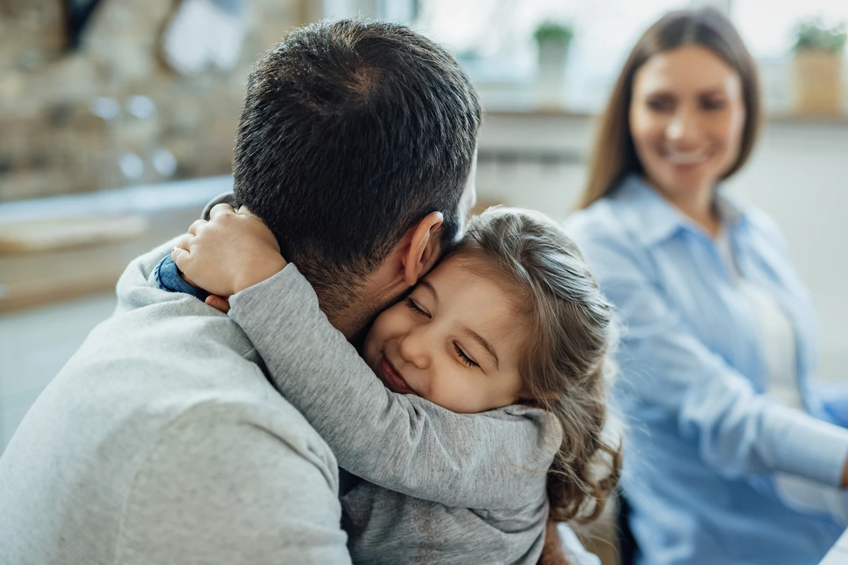 Affectionate little girl with eyes closed embracing her father at home. Mother is in the background. por Freepik