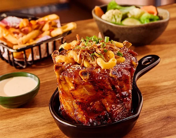 Ribs Rocker Crown: Costela suína servida em formato de coroa, recheada com macarrão cremoso com queijo, coberta com costela desfiada e acompanhada de batatas com queijo e um acompanhamento à escolha. por Divulgação